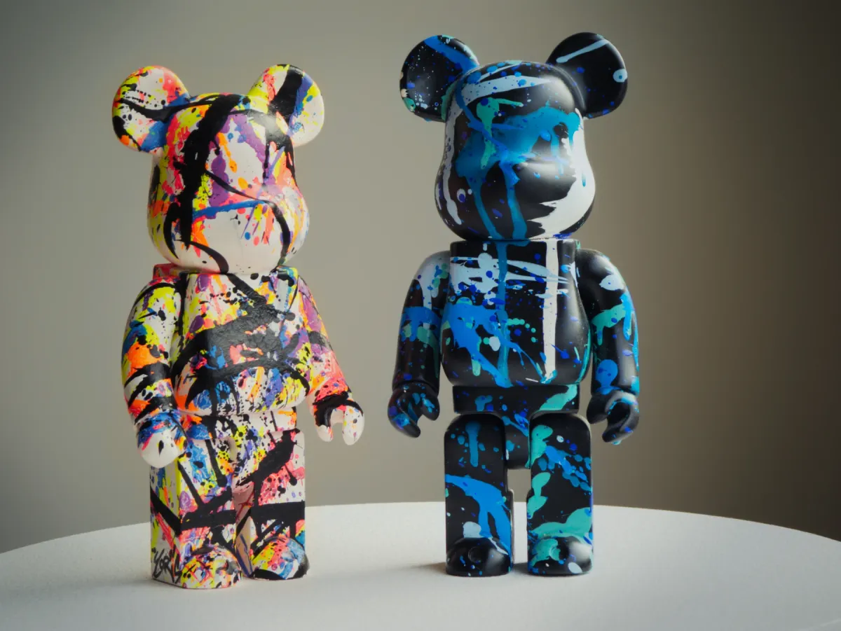 Deux figurines d’ours côte à côte sur un socle blanc : l’une éclaboussée de jaune, de rose et de bleu, l’autre noire striée de bleu et de blanc.