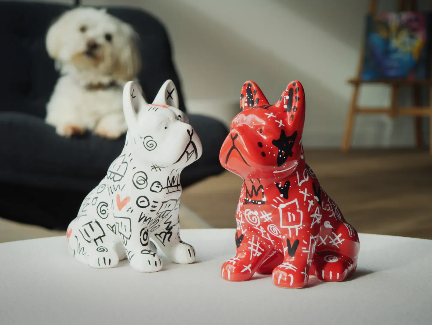 Deux sculptures de chiens, l’une blanche et l’autre rouge, sur une table ; un vrai chien blanc les observe depuis un fauteuil.