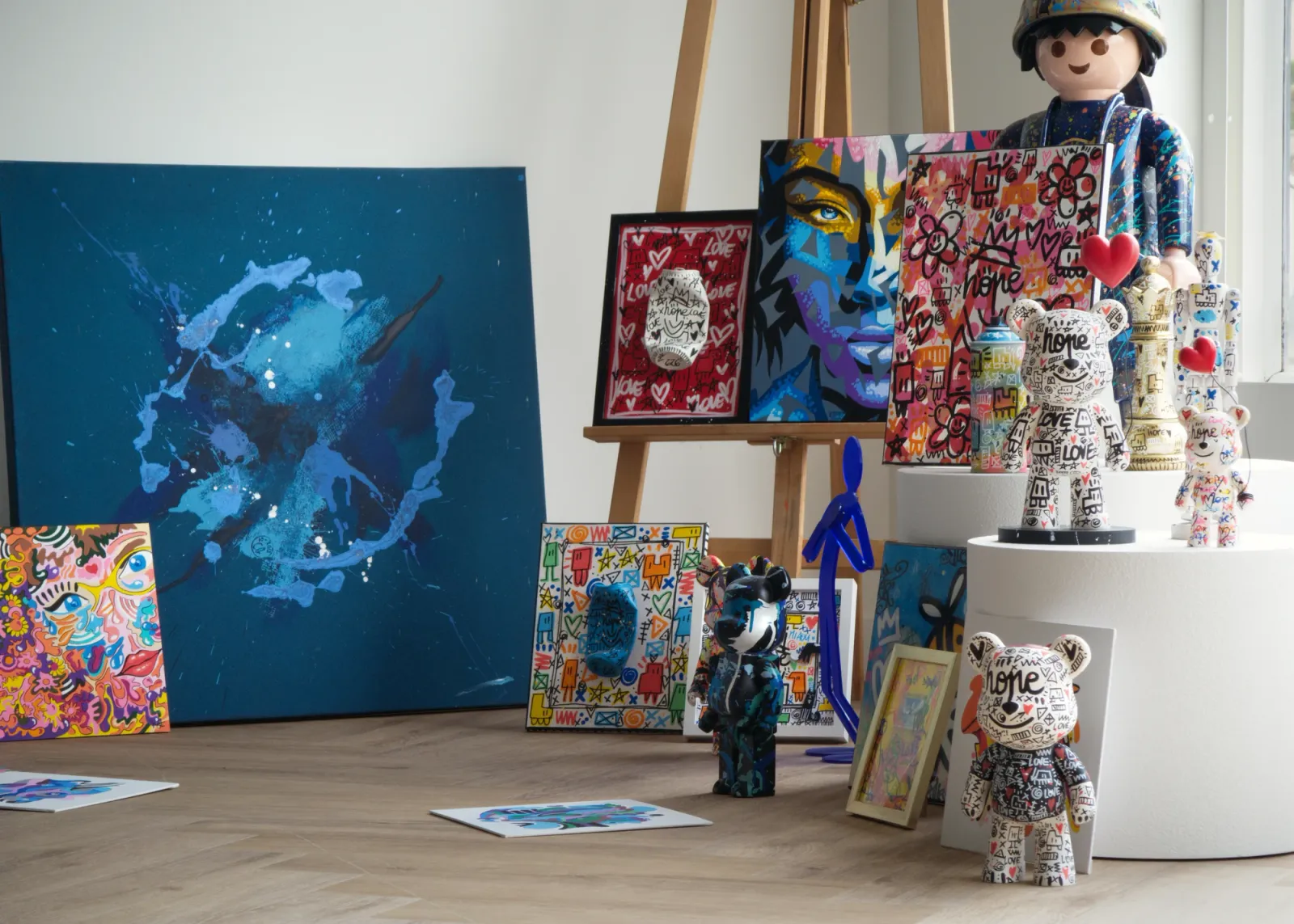 Peintures colorées, grande toile bleue et figurines réunies autour d’un chevalet, près d’une fenêtre.